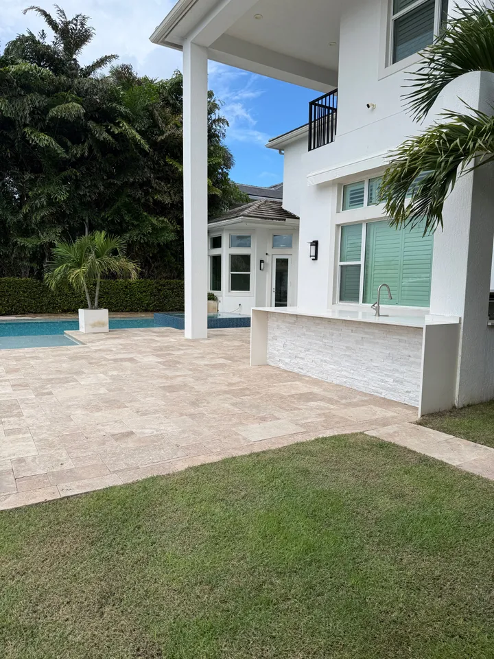 Clean travertine patio beside a pool and outdoor kitchen at a white two-story home